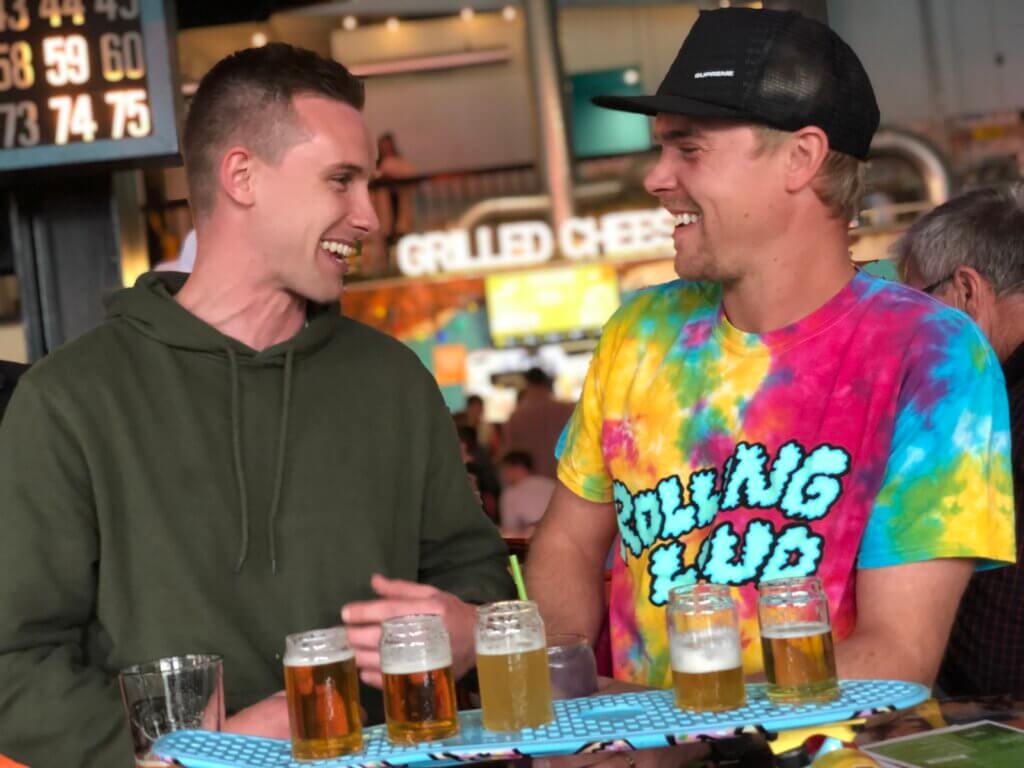 Two men laughing next to flight of beer served on a skateboard