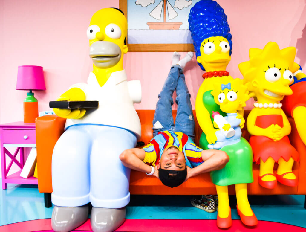 man laying upside down in bright colored clothes on couch next to models of the Simpsons