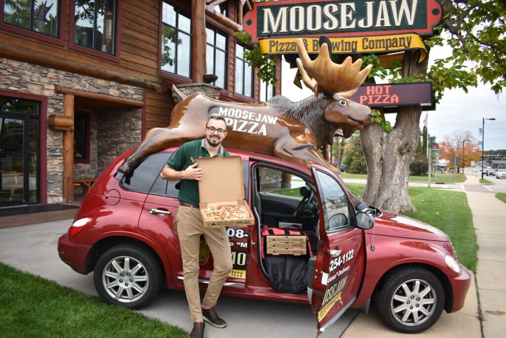 Moosejaw Pizza delivery driver holding a pizza next to a Moose-Mobile in front of restaurant