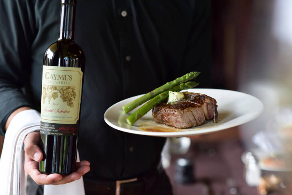 Server holding bottle of wine and plate of steak with asparagus for date night